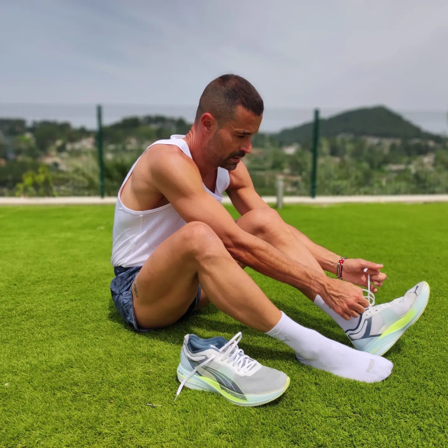 Un homme en tenue de sport est assis sur une pelouse en train de faire les lacets de ses chaussures PUMA