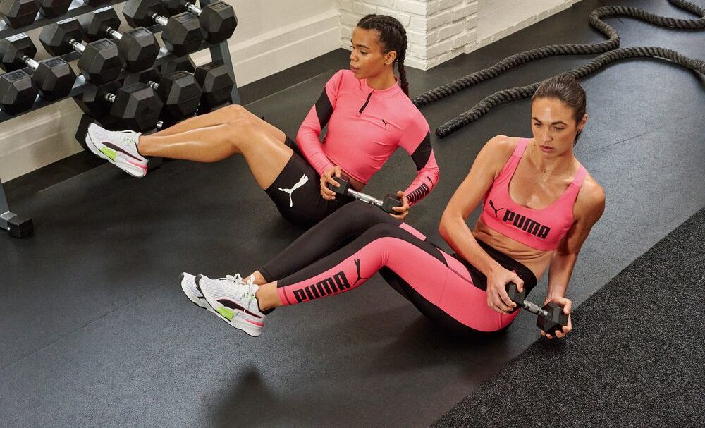 Deux femmes en tenues de sport PUMA roses font des exercices d’abdominaux dans une salle de fitness