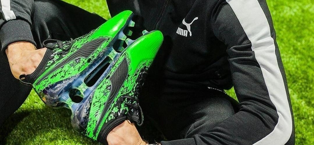 Close-up van iemand die op een voetbalveld zit en een paar groene PUMA voetbalschoenen met de noppen tegen elkaar in zijn handen houdt.