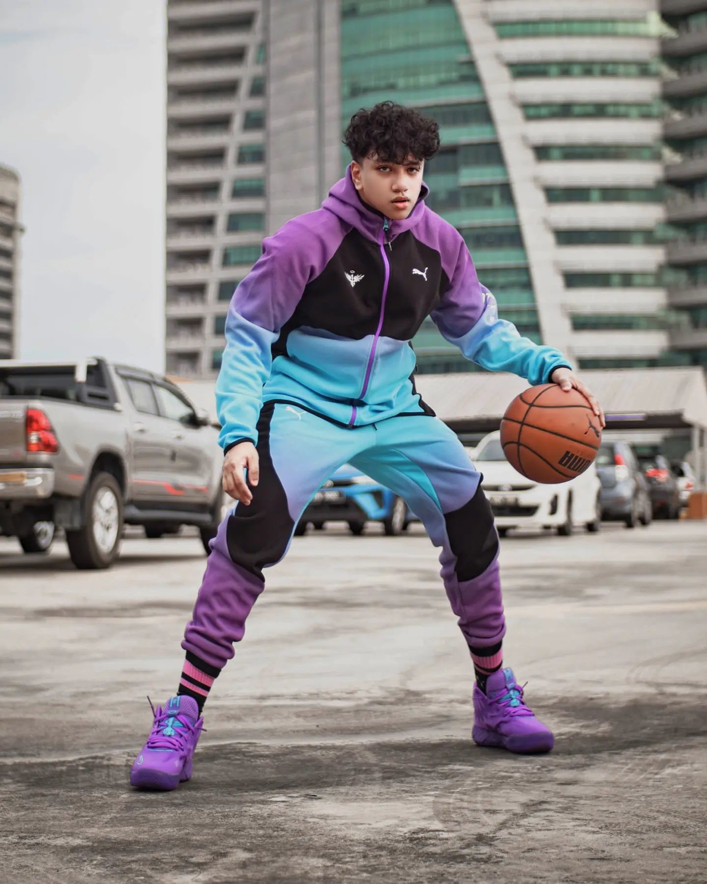 Basketballspieler in schwarz-blau-violetem PUMA Outfit und lilanen Baskettballschuhen beim Dribbeln am Parkplatz