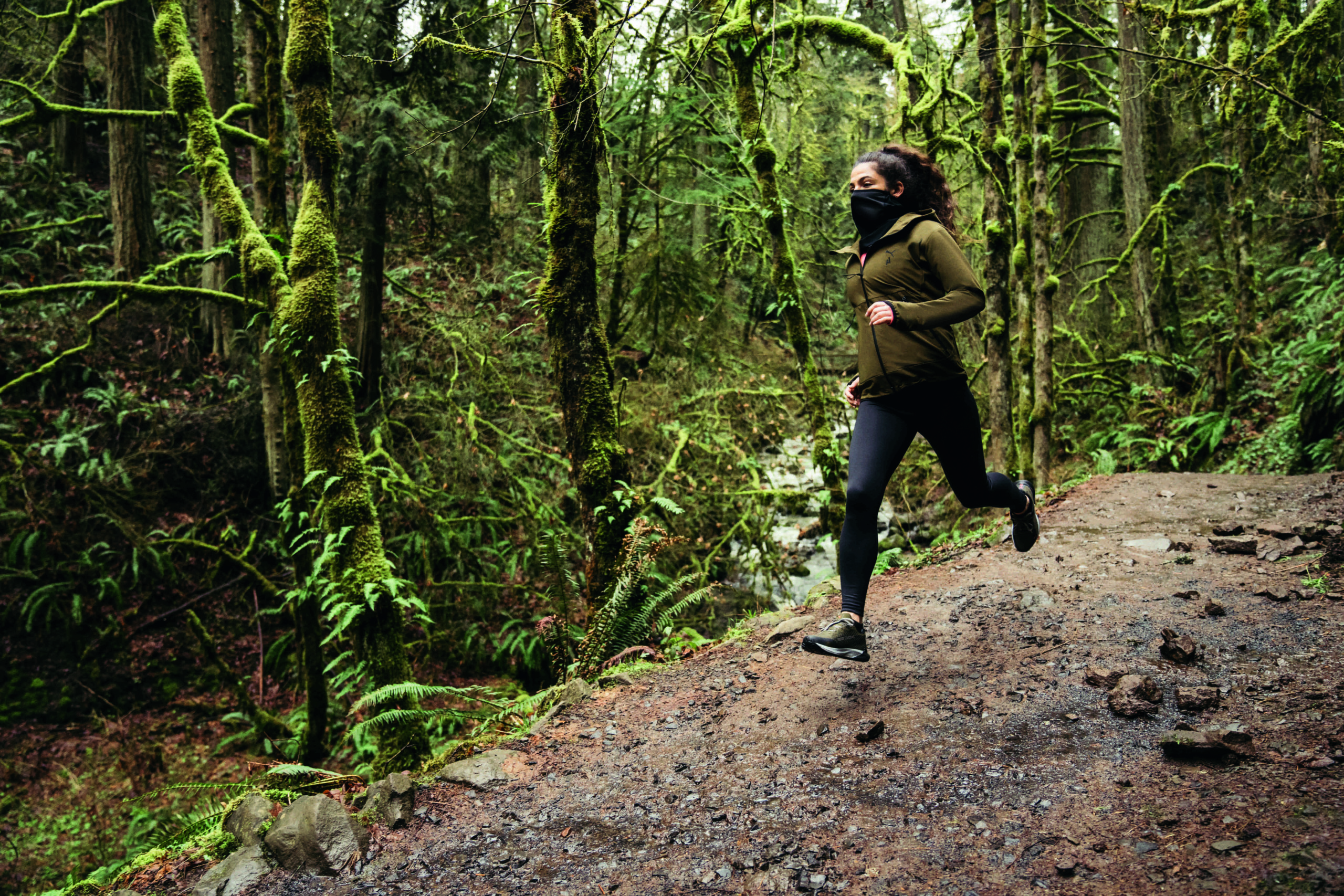 Läuferin beim Trailrunning im Wald gekleidet in schwarzen PUMA Laufleggins, einer olivgrünen PUMA Laufjacke und mit Fast Trac Nitro GTX Trailrunningschuhen in olivgrün-schwarz