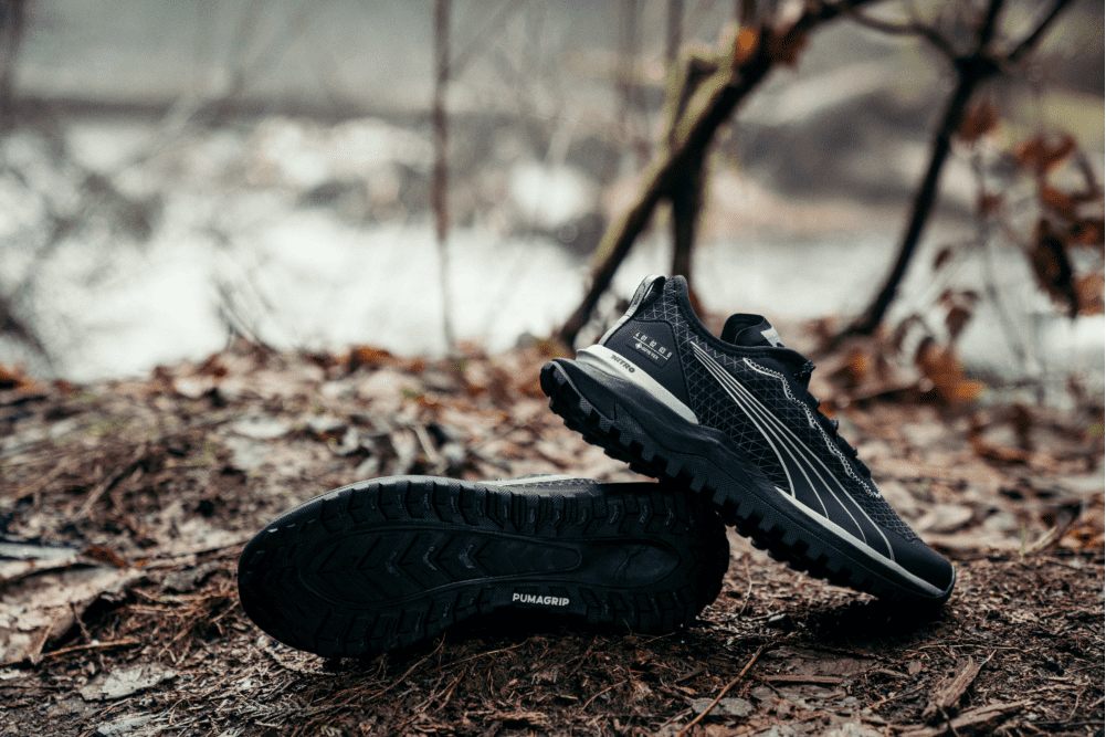 Een zwart paar trailschoenen in het bos waarvan je de onderkant ziet van een van de schoenen