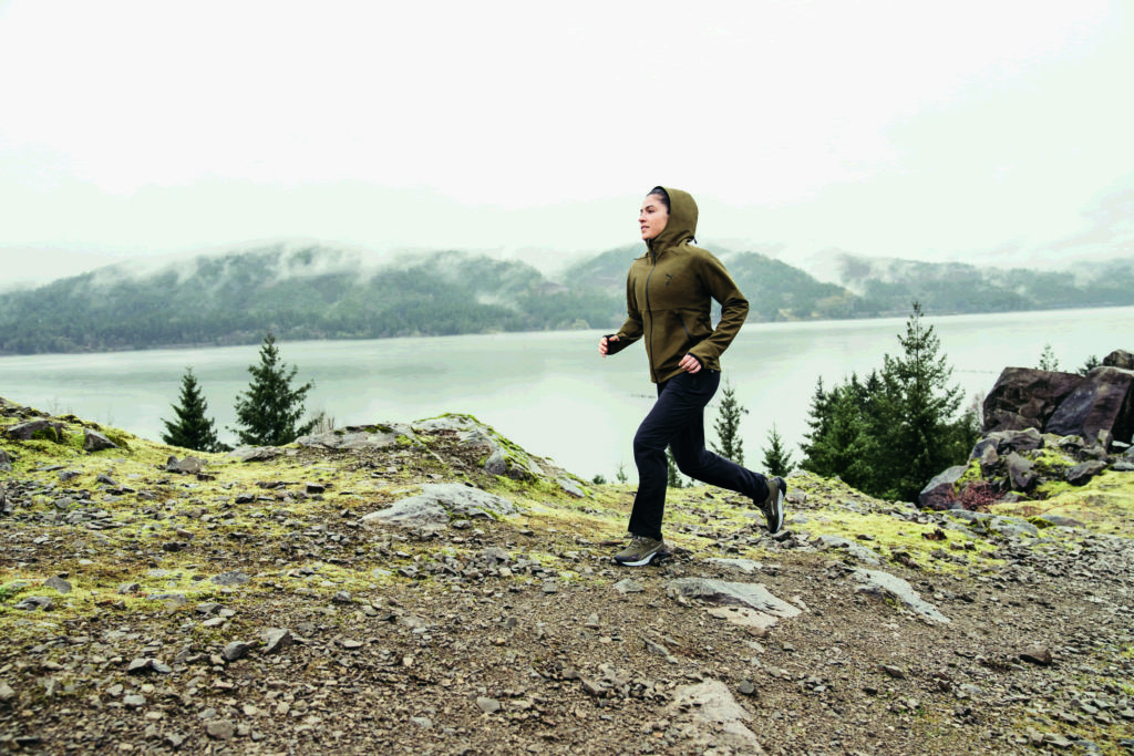 Une femme en tenue de trail PUMA court en montagne devant un lac 