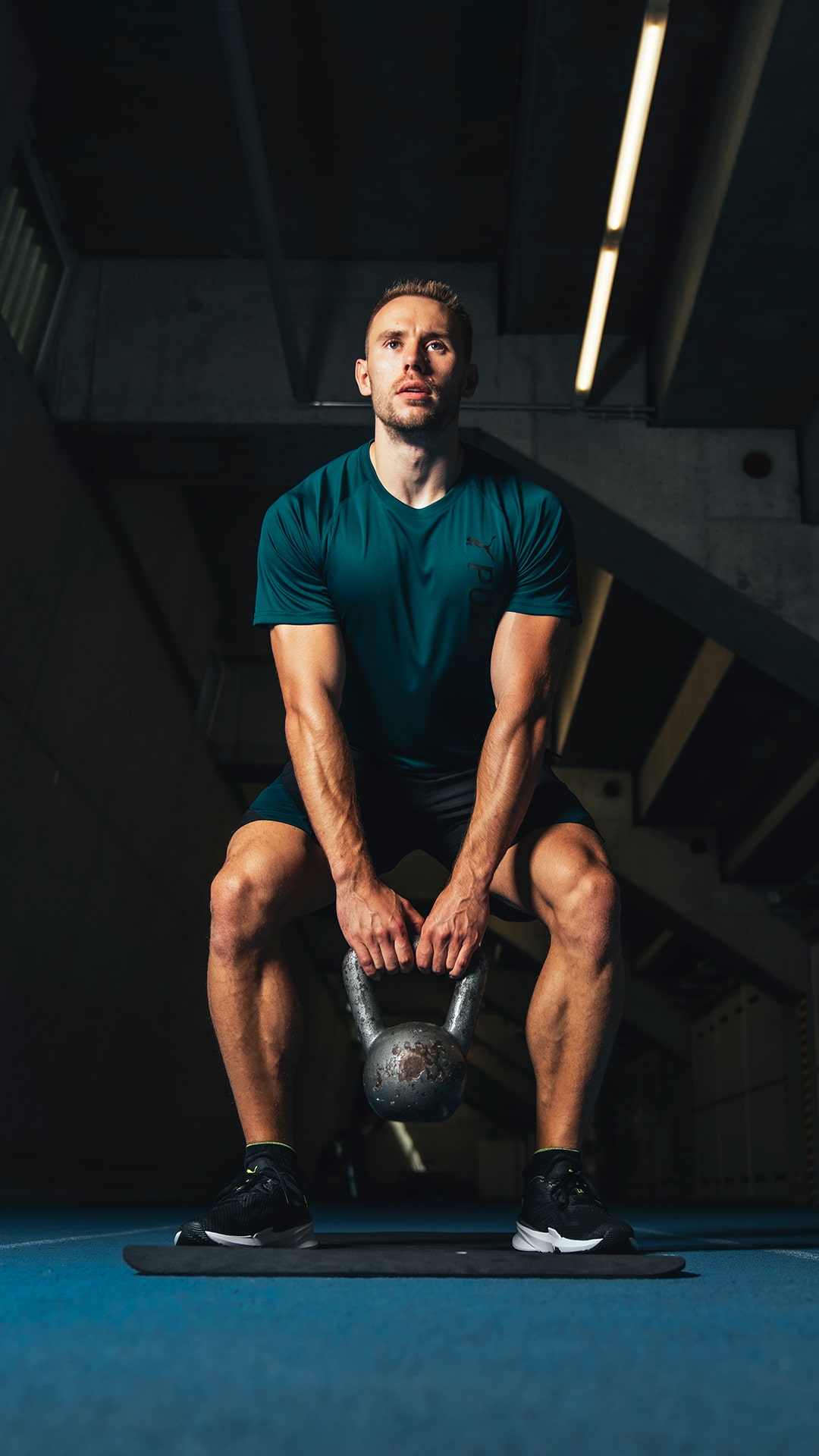 Athlet in petrolfarbenem PUMA Shirt und Shorts macht Kniebeugen mit Kettlebell im Fitnessstudio