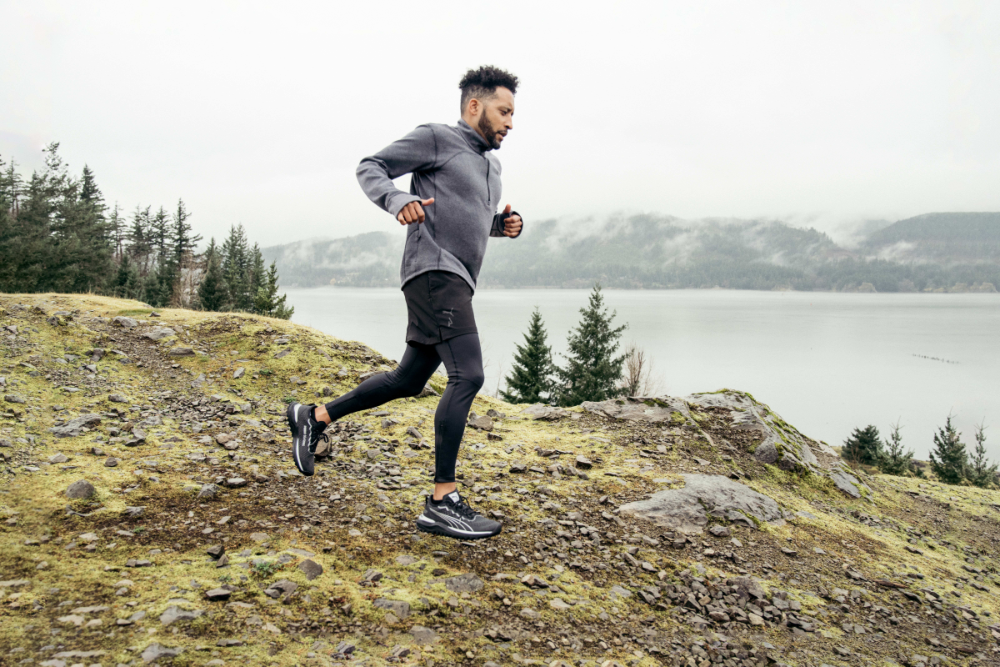 Läufer beim Trailrunning in hügeligem Wald gekleidet in schwarzer enger Laufhose, schwarzen Laufshorts und einer grauen Laufjacke von PUMA