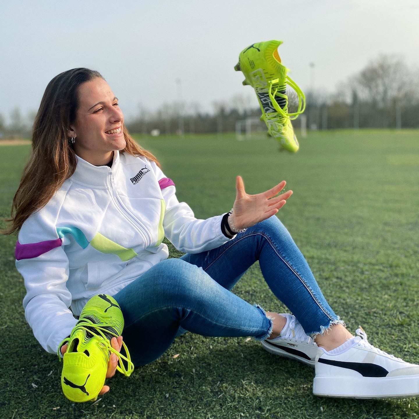 Spanische Fußballerin Laia Codina in Jeans und weißer PUMA Sportjacke sitzt auf Rasen und wirft ihre neongelben PUMA Fußballschuhe in die Luft