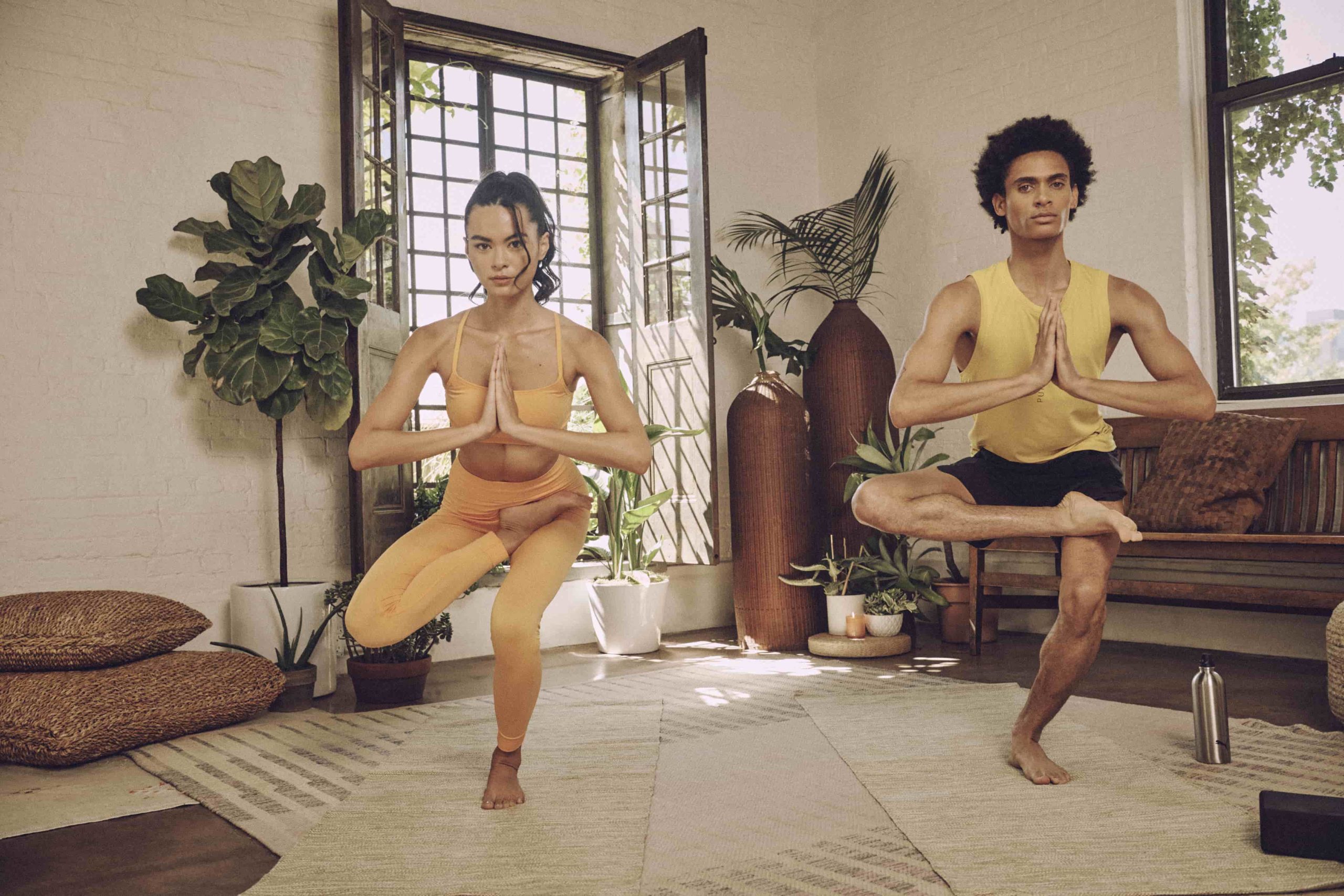 Frau in orangenem Yoga Set und Mann in kurzer schwarzer Hose und gelbem Tank Top in Eka Pada Utkatasana Yoga Stellung